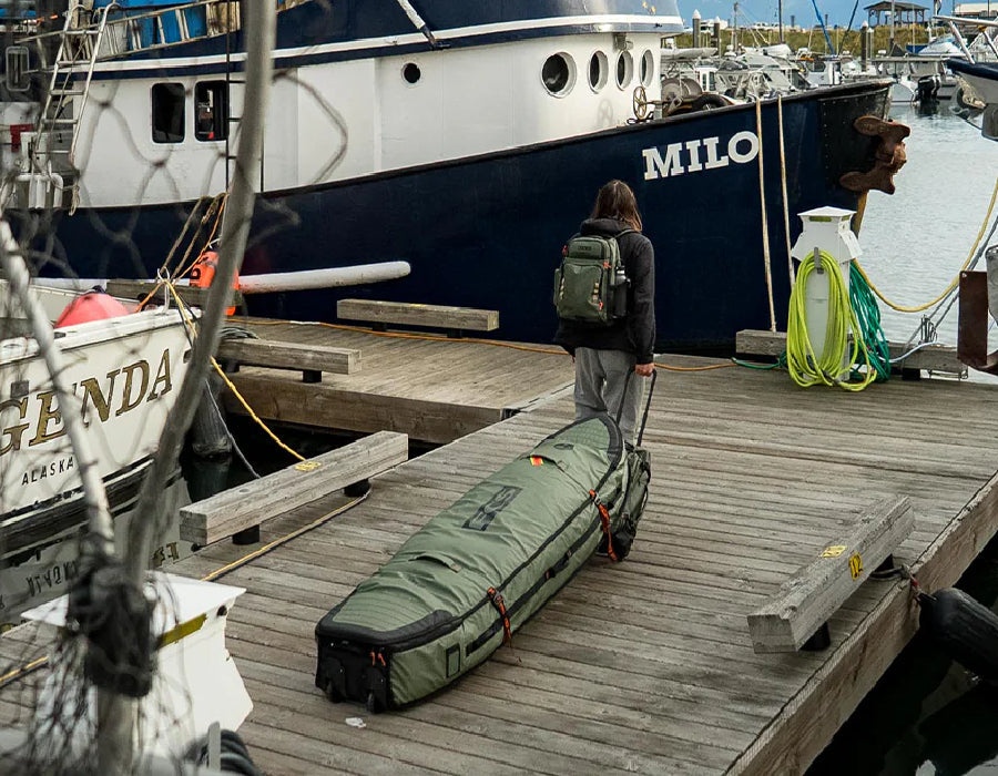 Person with backpack rolling surfboard travel bag on dock near boats, Melbourne Surfboard Shop