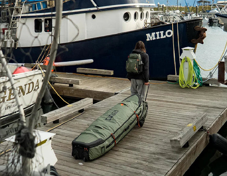 Person with backpack rolling surfboard travel bag on dock near boats, Melbourne Surfboard Shop