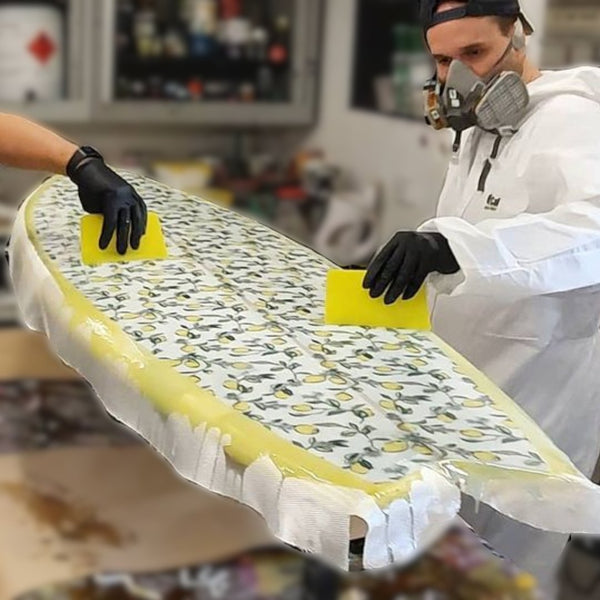 Glassing a surfboard by applying resin over fibreglass cloth with squeegees.
