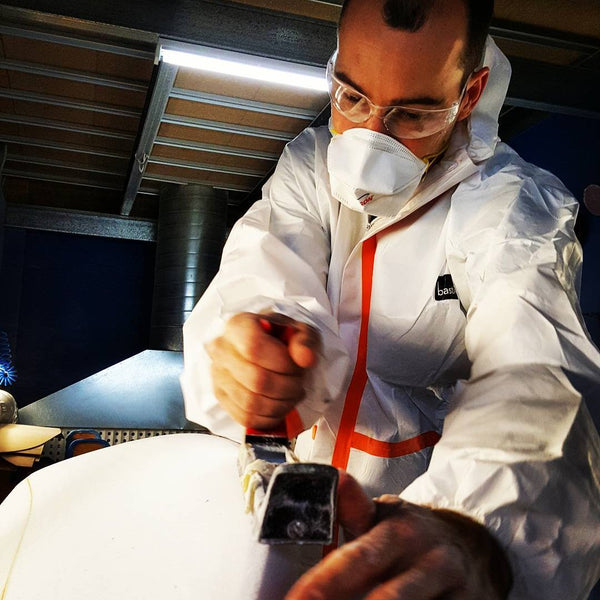 Sanding down a surfboard blank with protective glasses, mask and coveralls.